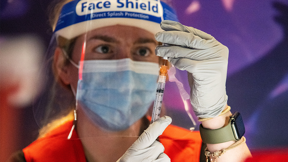 An individual wearing gloves, a mask and face shield prepares a vaccine dose in a syringe