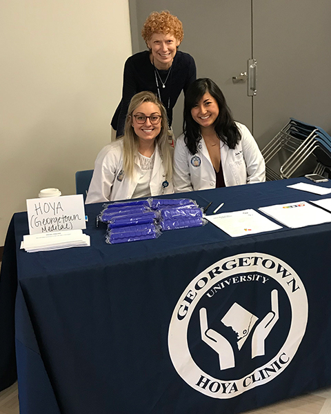 Three women stand together behind a table promoting HOYA Clinic at a health fair