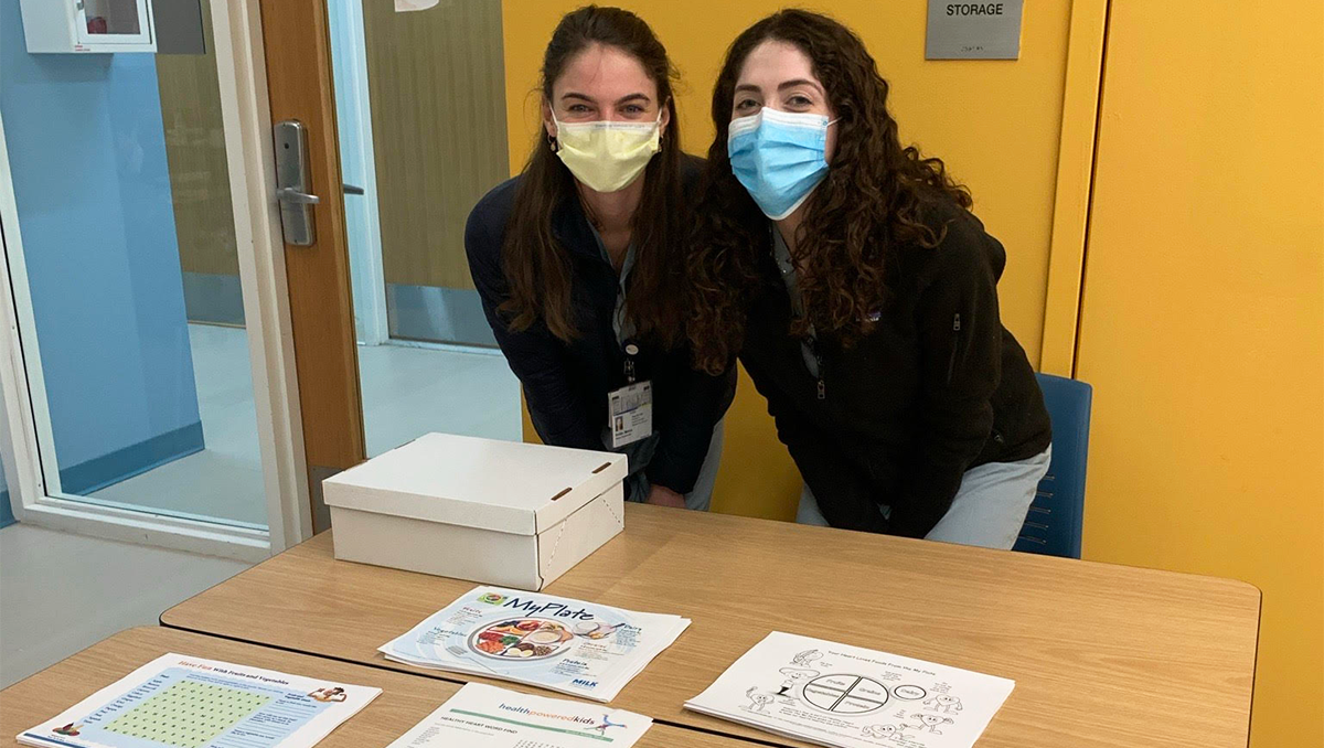 Two students wearing masks stand side by side behind a table filled with paper fliers