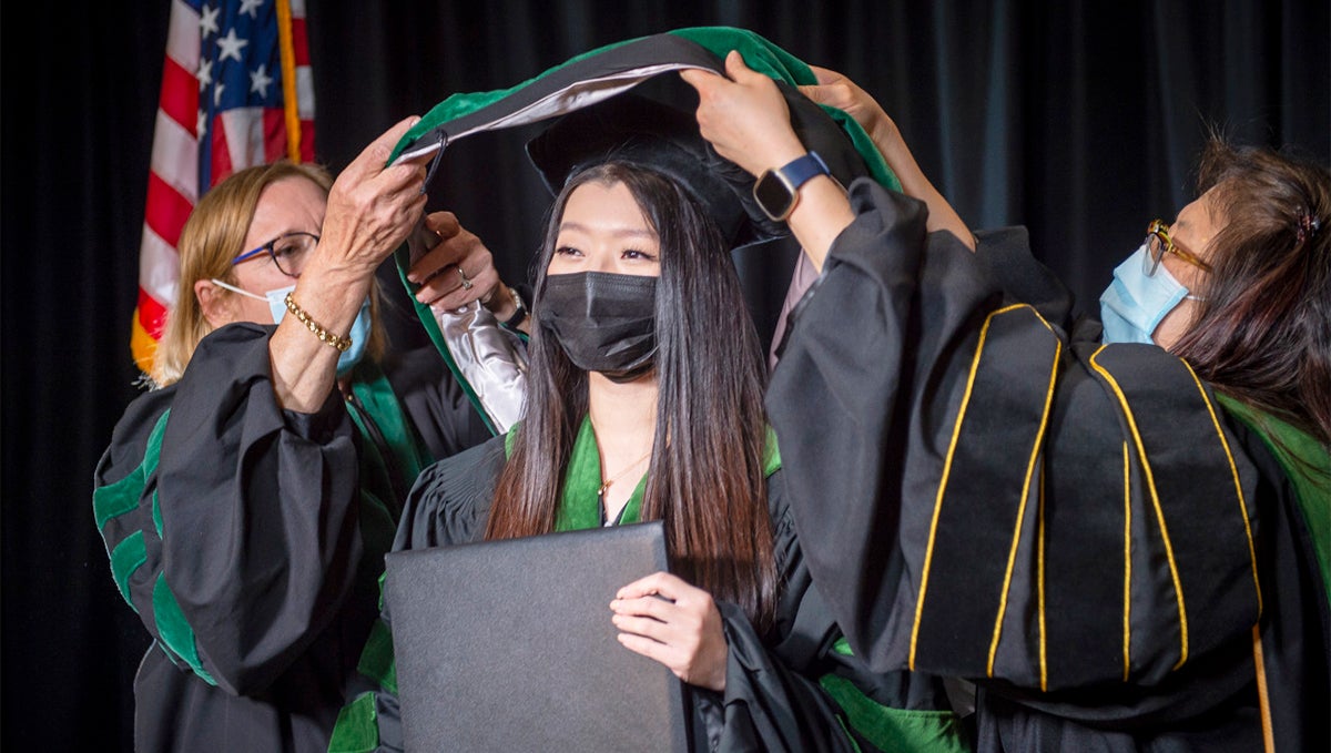 Two women lift a ceremonial hood over the shoulders of a third woman in academic regalia