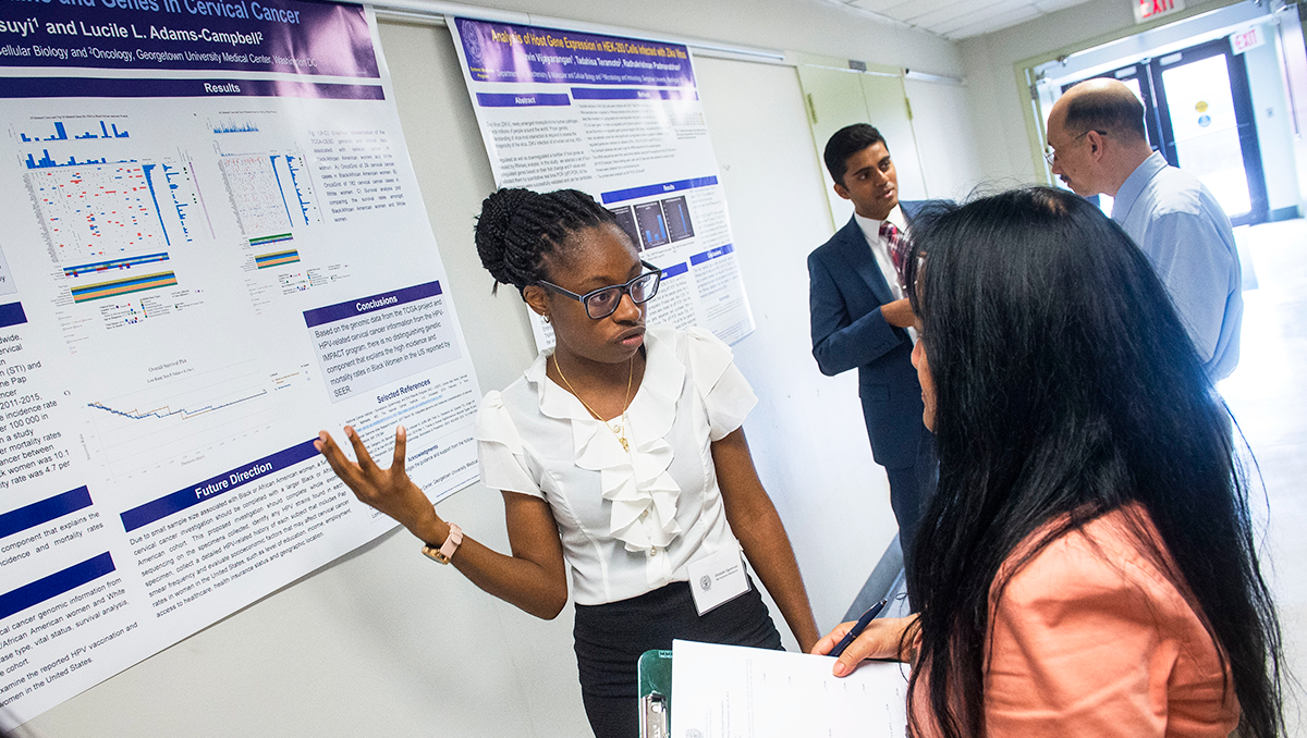 A student gestures toward a poster on the wall while speaking with a professor