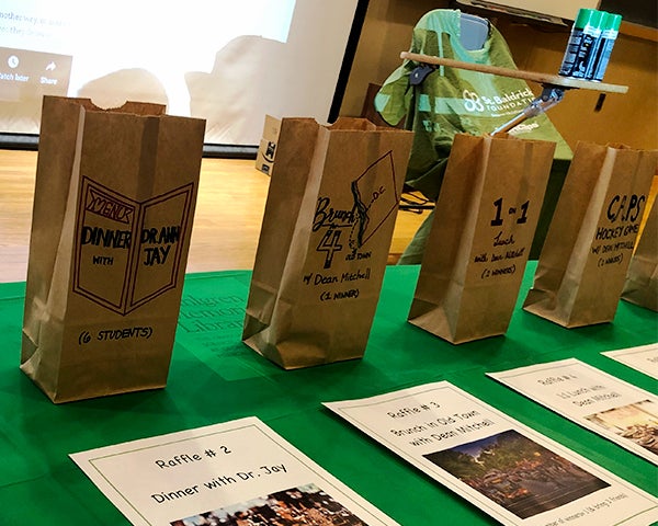 Decorated paper bags to hold tickets line a table filled with signs for each raffle item. Behind them is a stage with a desk draped with a St. Baldrick's barber's cape.