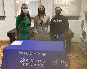 Three individuals, including Dr. Seliby Perkins, stand indoors behind a table draped with a tablecloth printed with logos for MIECHV, Mary's Center, and DC Health 