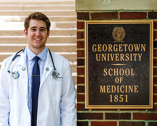 Brian Fitzgerald pictured in his white coat with stethoscope next to the Georgetown University School of Medicine sign outside the Med-Dent building