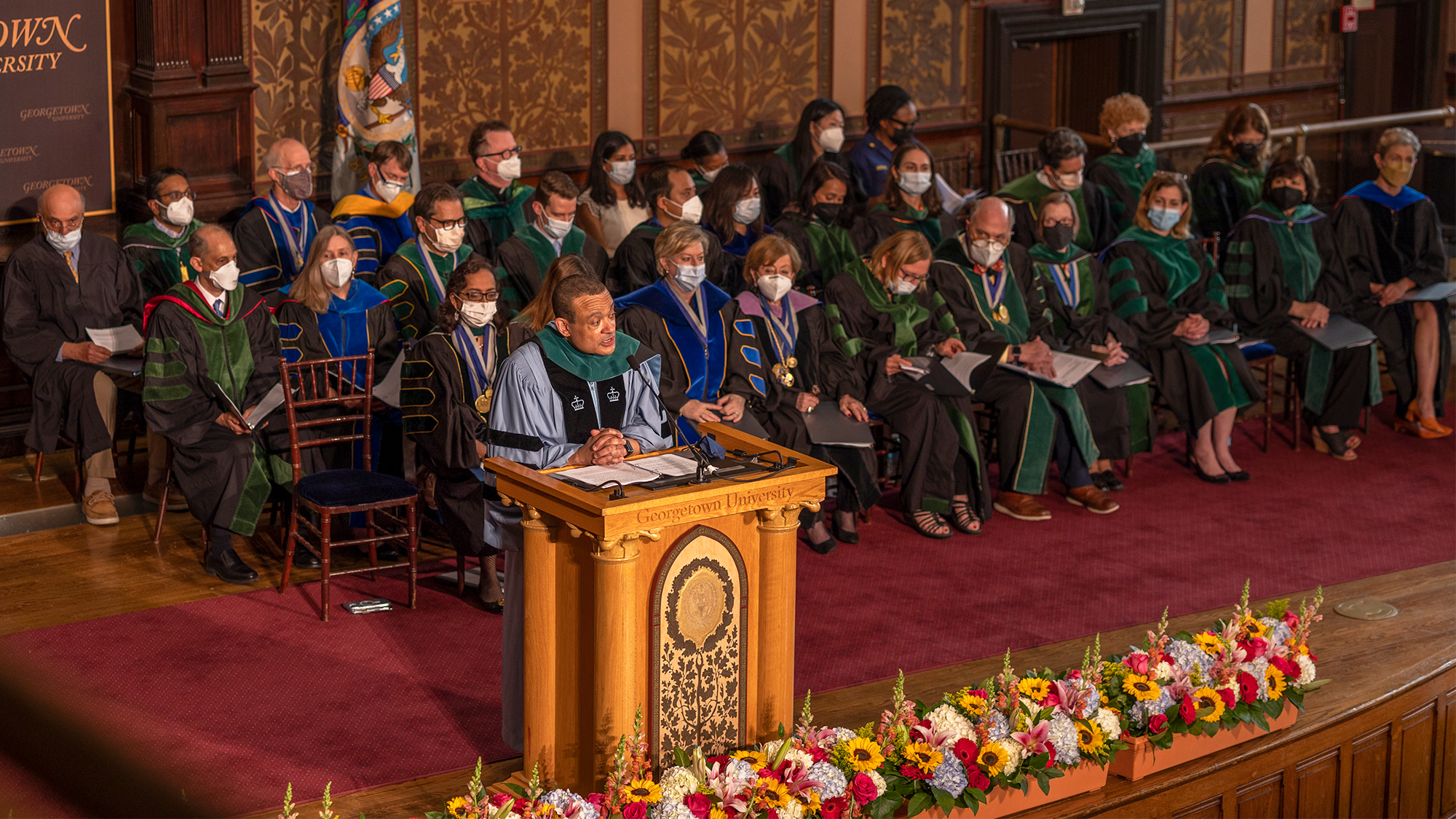 Dean Jones in commencement regalia speaks from a podium on a stage where faculty members are seated