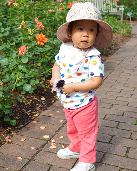 A tiny girl wearing a hat stands outside next to roses