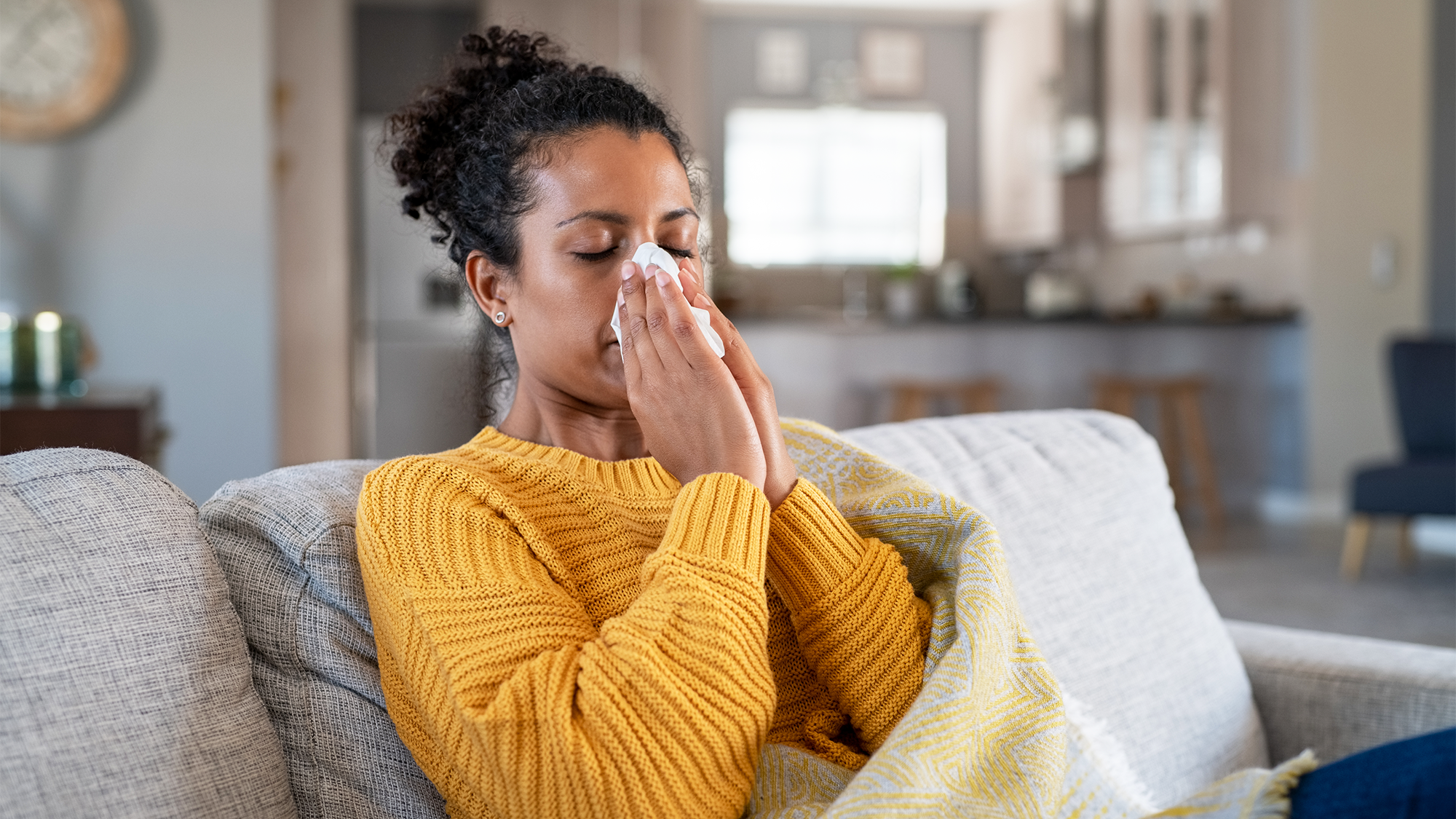 A woman blows her nose while sitting on a couch wrapped in a blanket obviously ill