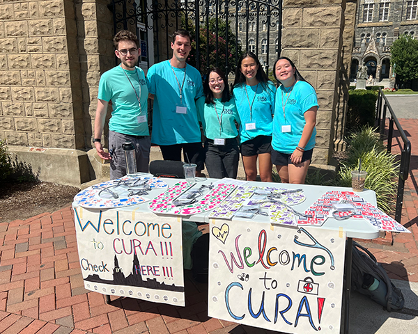 Five students stand a group behind a table adorned with signs that say Welcome to CURA!