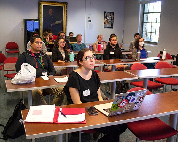 Students sit at tables with attention focused at the front of the room