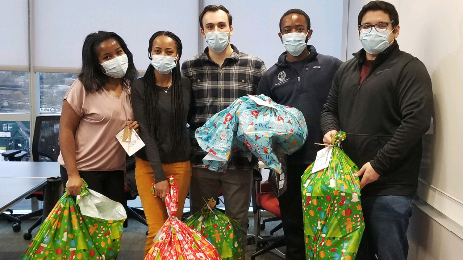 Five students stand together holding large, festive holiday bags filled with gifts