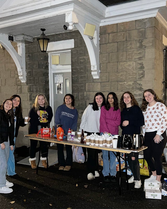 Eight students stand around a table outside filled with cups of hot chocolate