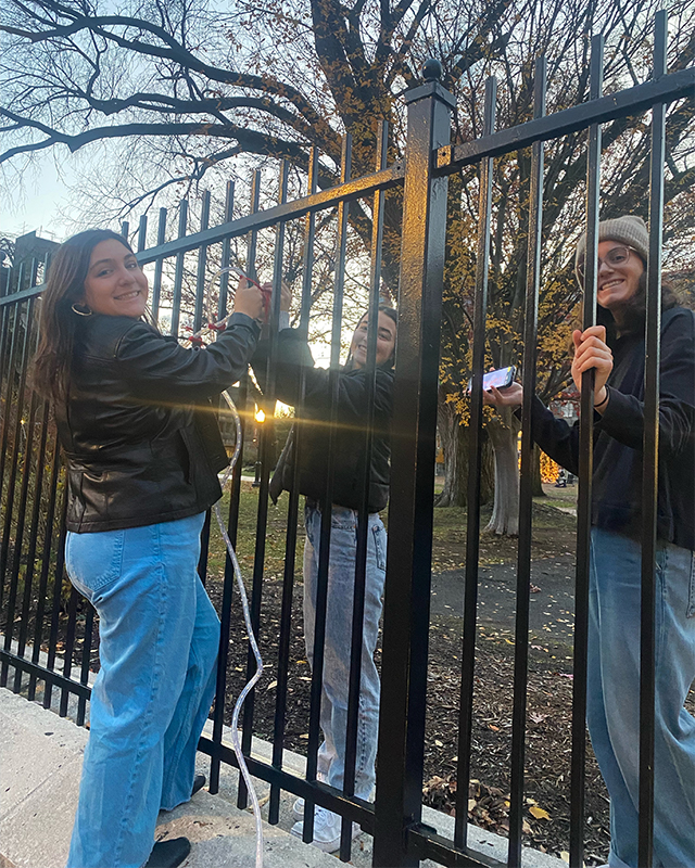 Three students decorate a fence with holiday lights