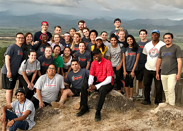 A large group of students stand around dean Brian Floyd with a view of a plain and mountains in the background