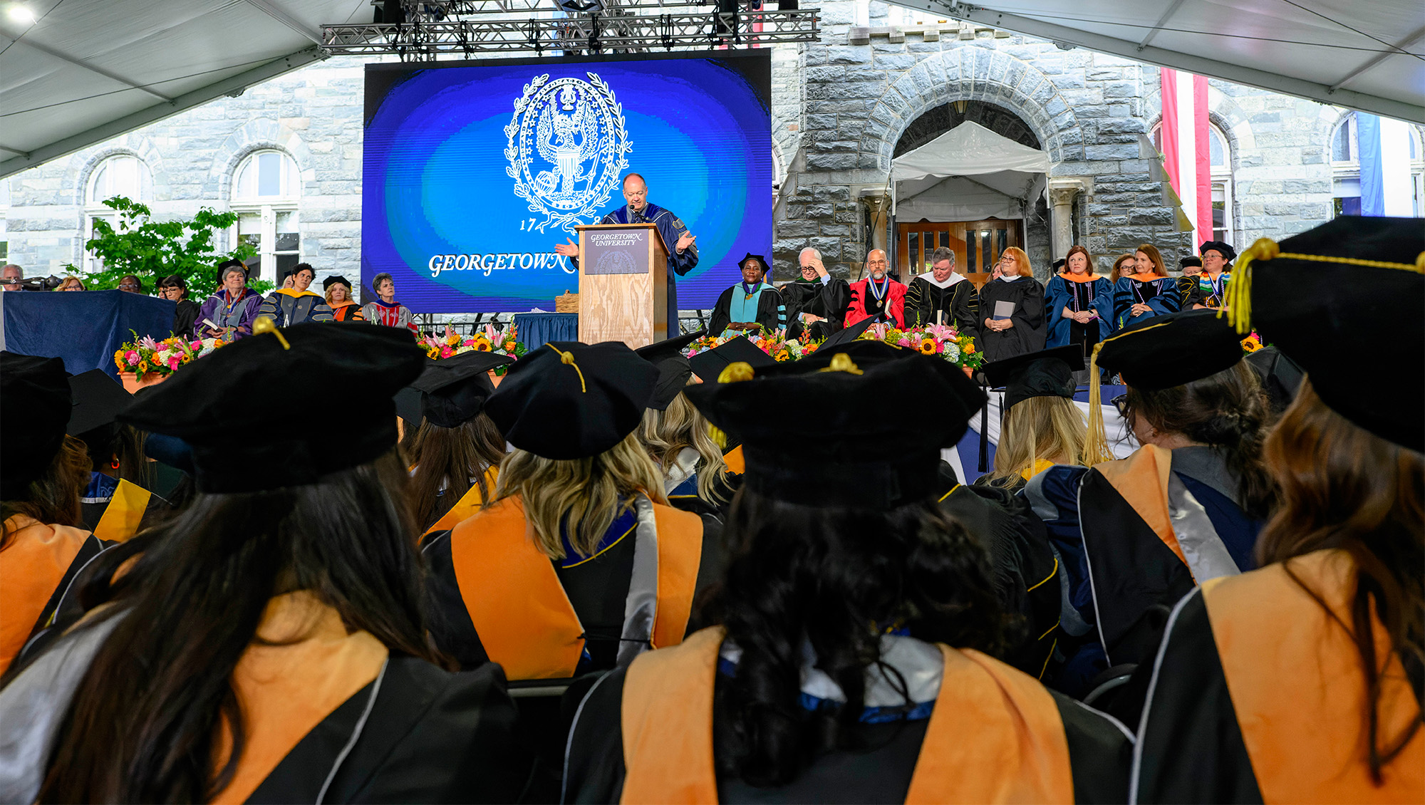 President DeGioia stands at a podium during Commencement looking out at an audience of School of Nursing graduates, behind him are seated school faculty