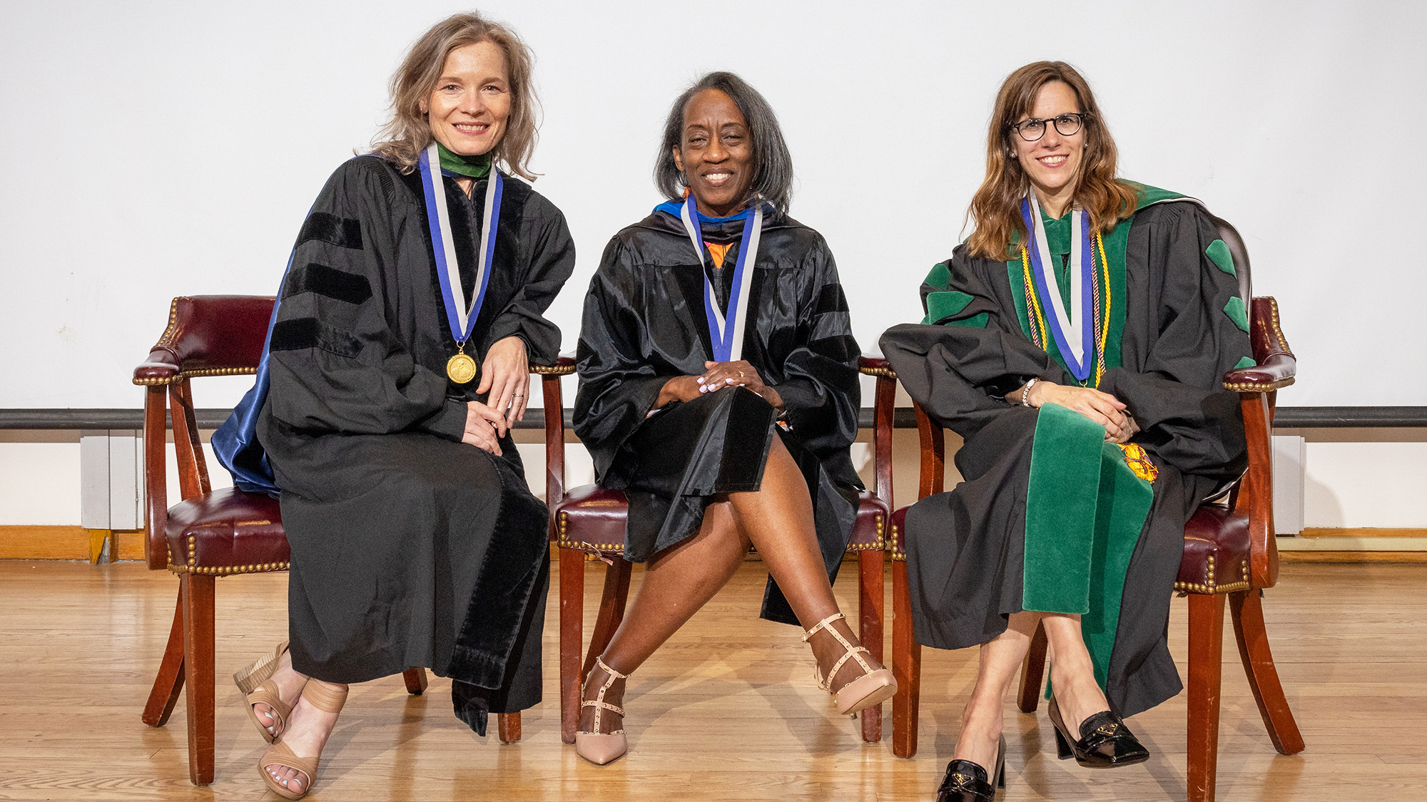 Three women dressed their academic regalia sit in chairs