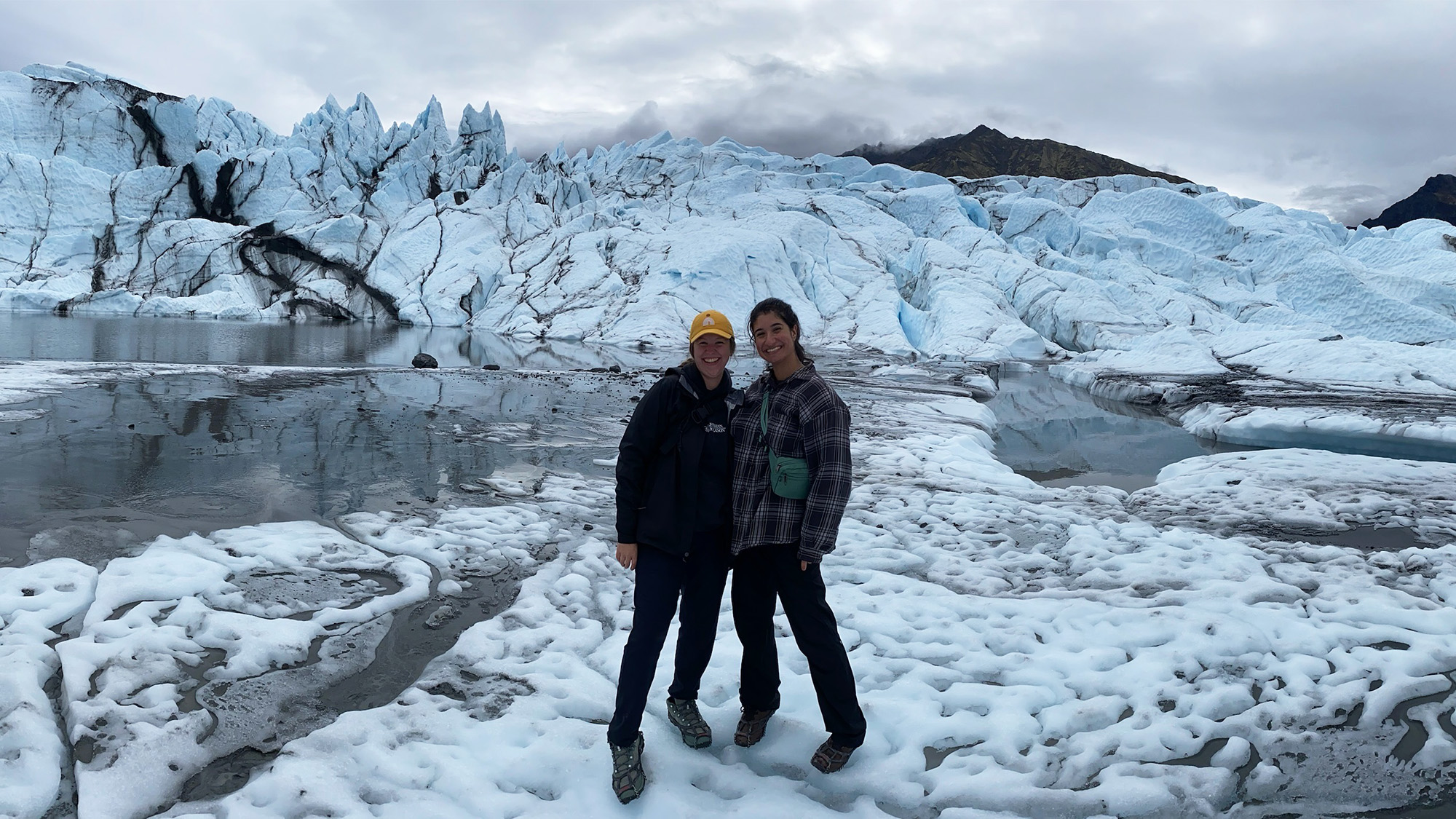 Two people stand near a glacial lake with a glacier in the background