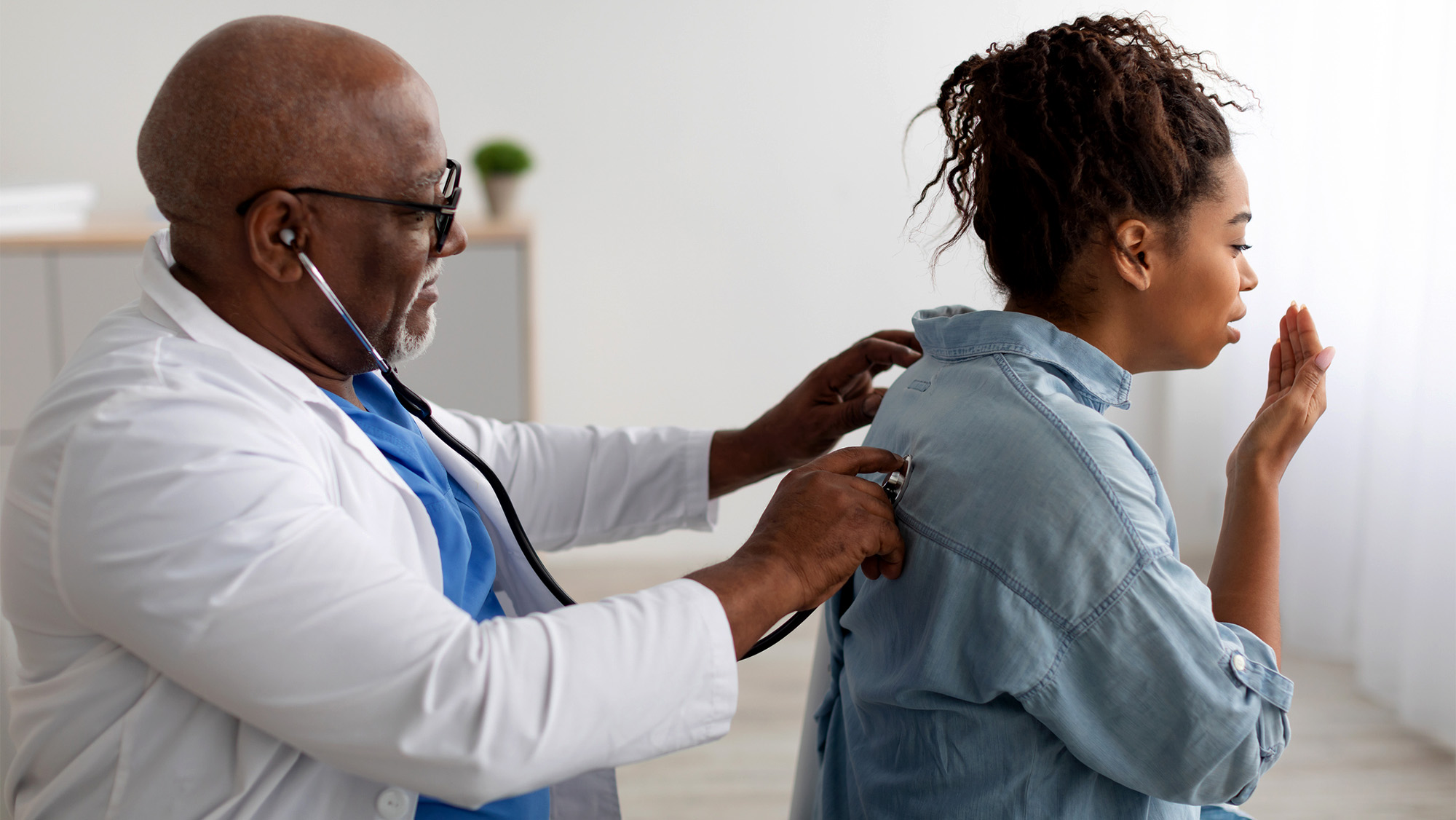 A doctor holds a stethoscope to a patient's back as she coughs