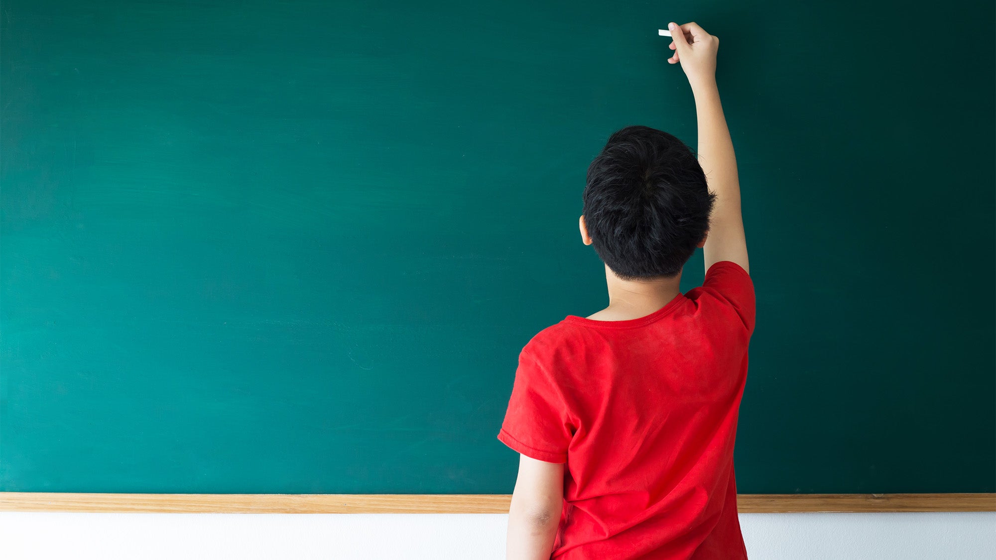 A boy stands at a blank chalkboard holding a piece of chalk