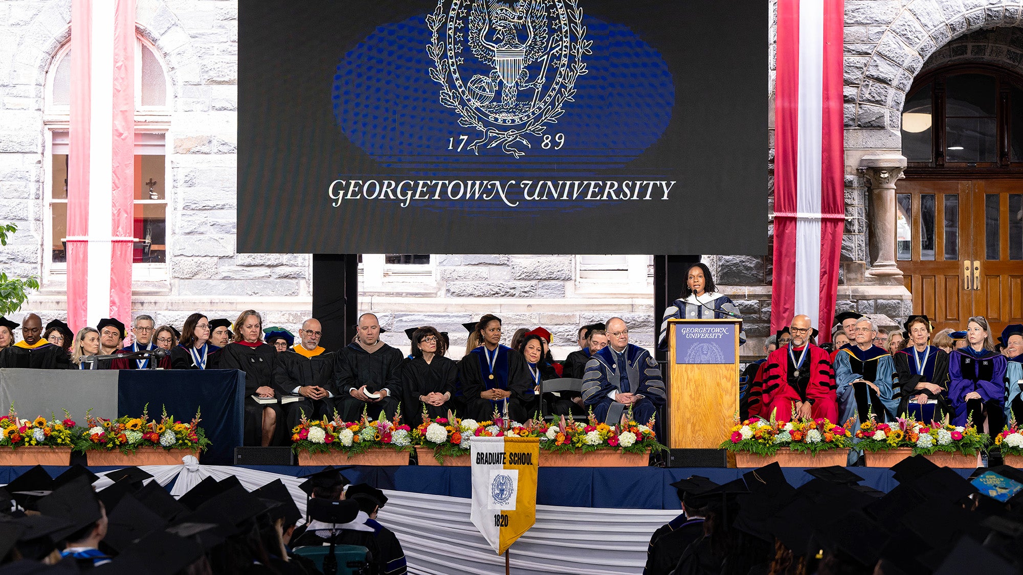 A wide view of the stage at Commencement where the speaker is talking at a podium and the faculty are seated behind her