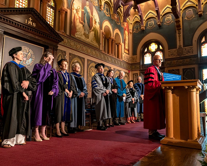 Faculty stand behind Alex Sens as he speaks from a podium at the hooding ceremony
