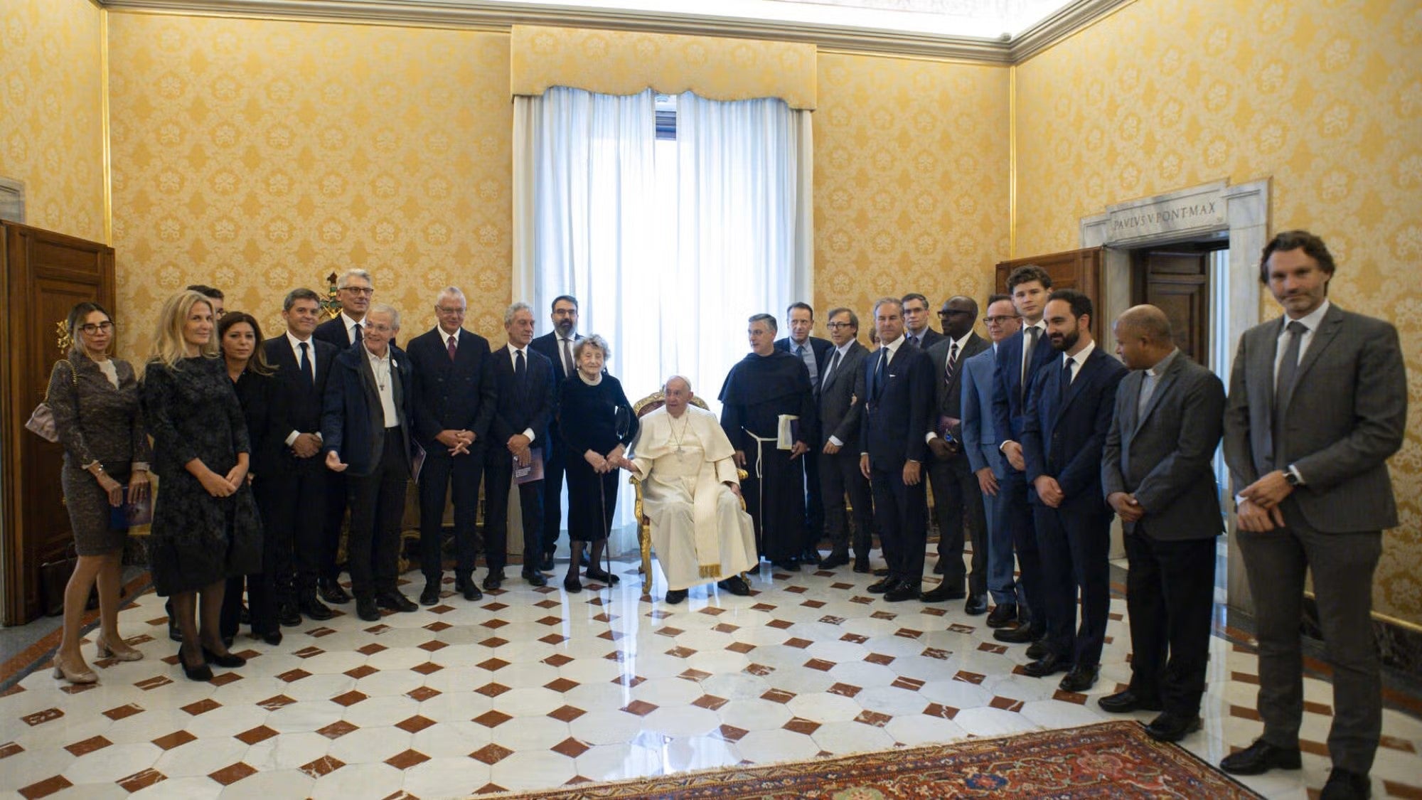 A large group of people stand around Pope Francis, who is seated in a chair, in an ornate room at the Vatican