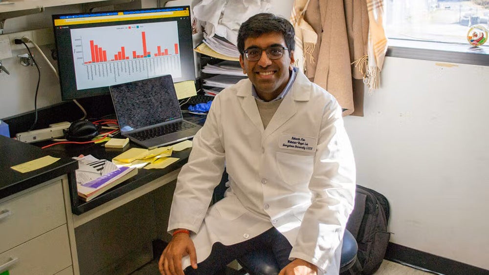 Sidharth Jain wears a white lab coat and sits in front of a computer in a lab setting