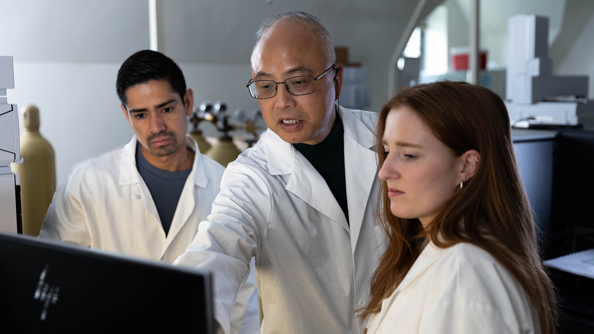 Two students and a professor in white coats look at a computer screen together