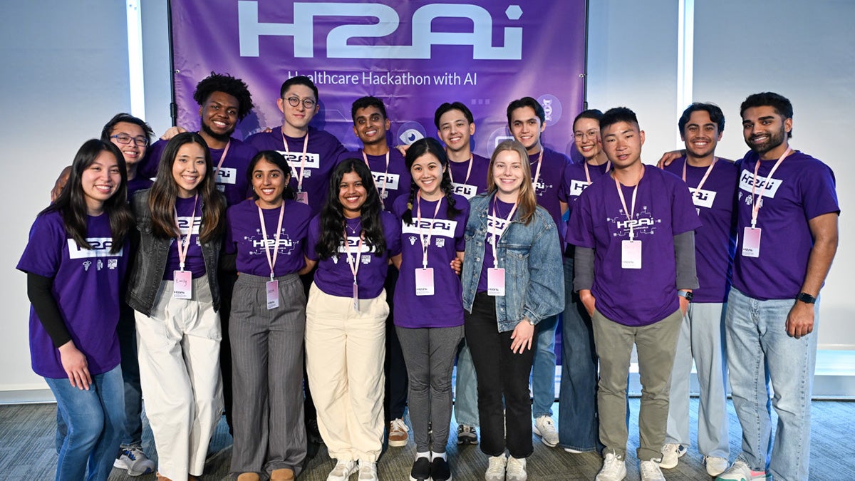 The student organizers of the H2AI event wear matching purple t-shirts and stand together in front of a banner for the event