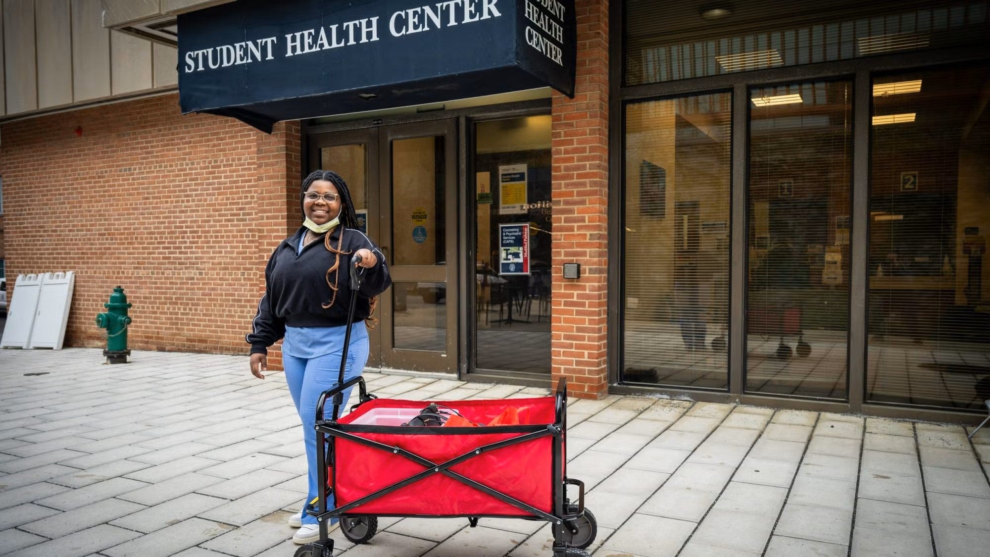 A Black woman stands with a red wagon in front of the Student Health Center on campus