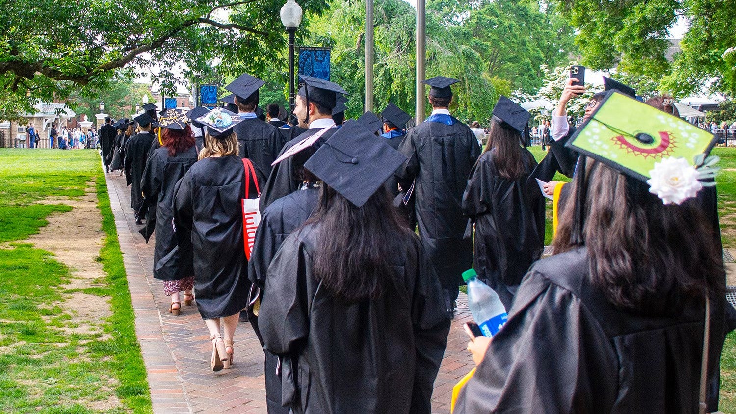 A line of students in caps and gowns walks down a sidewalk on campus