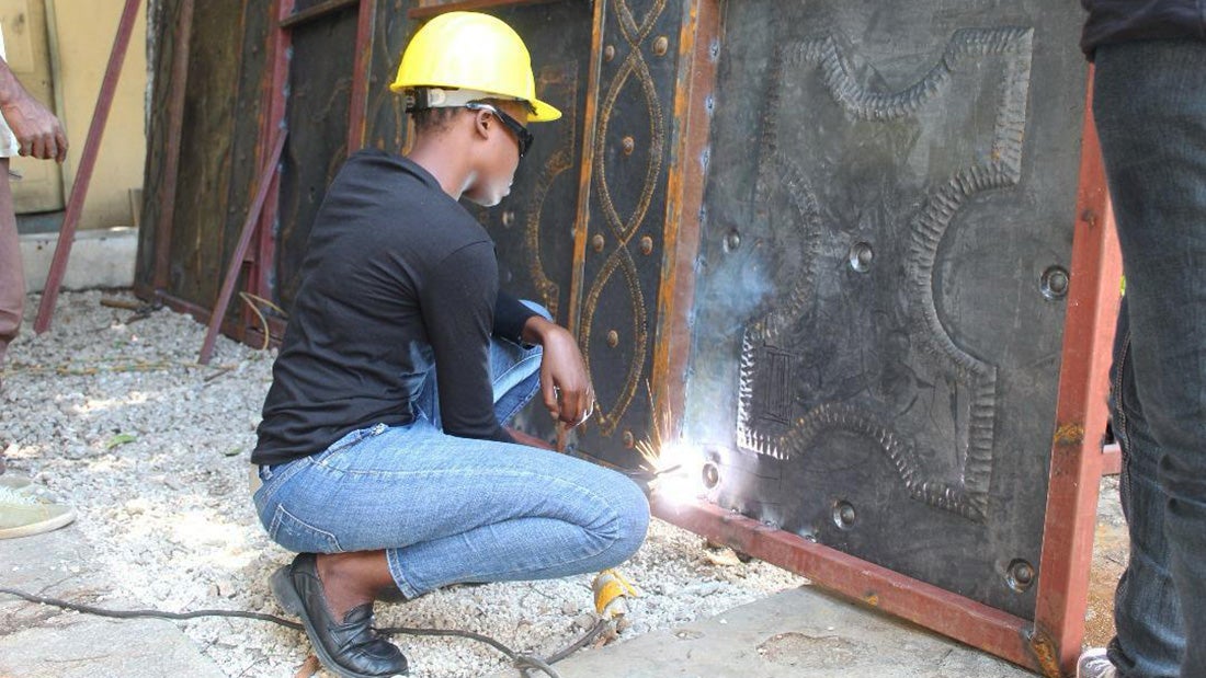 A young Haitian woman wearing safety equipment welds a decorative metal piece
