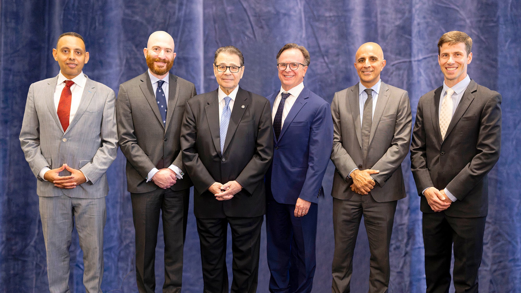 Six individuals stand together against a blue cloth backdrop