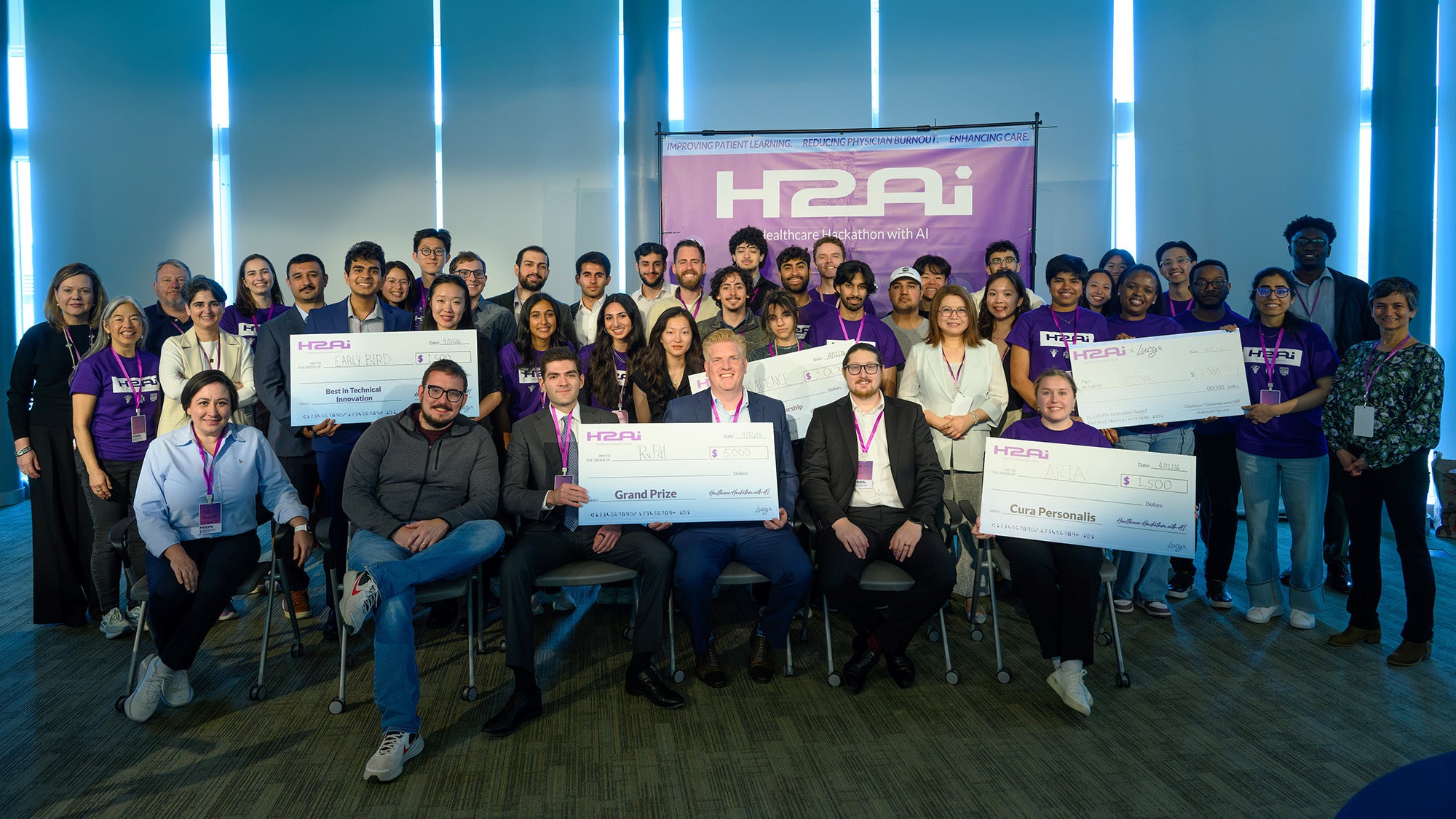 The participants in this year's H2AI Hackathon stand together in a large group with winning groups holding their large cardboard check awards