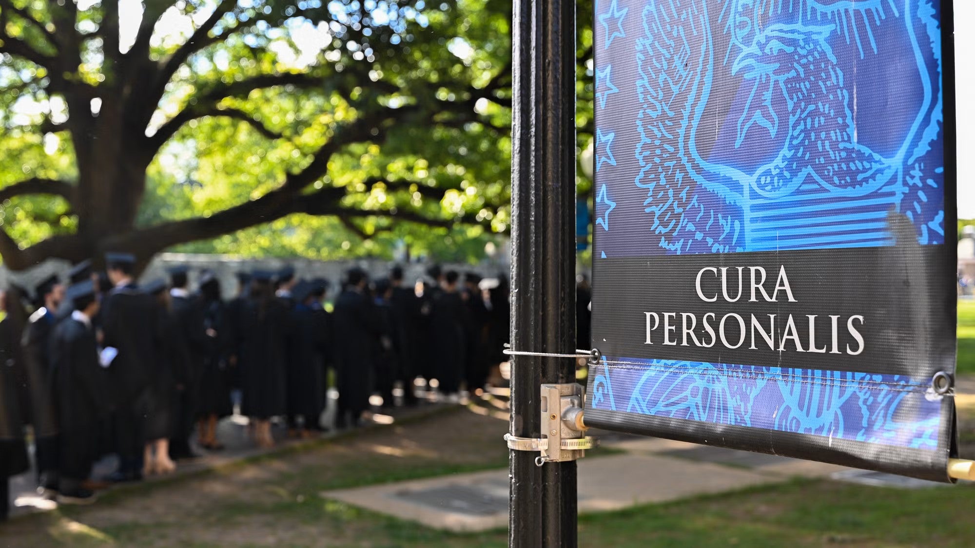 Graduating students process by a banner bearing the words Cura Personalis