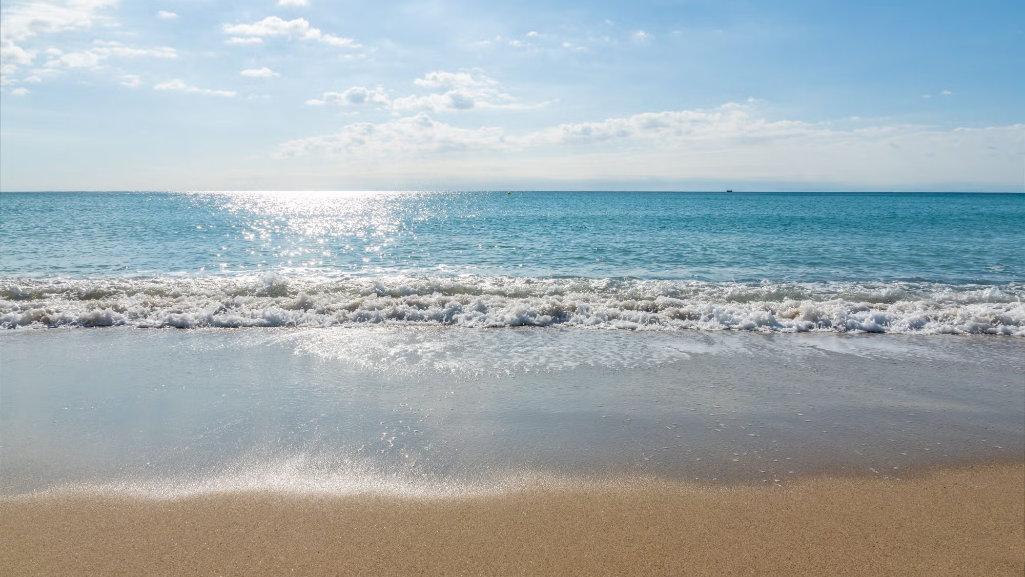 Ocean waves roll onto a beach over which the sun shines brightly