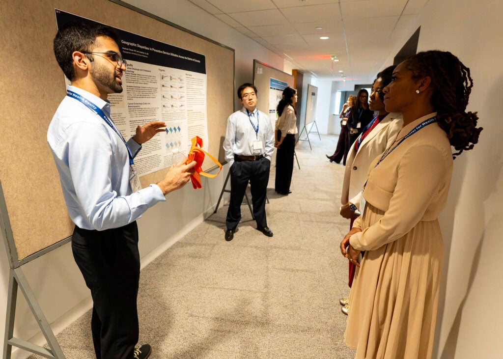 Two individuals present their poster to several others in a hallway area